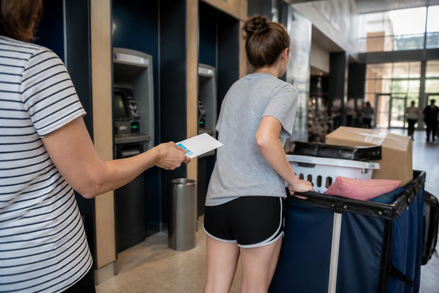 A parent handing a card sleeve to a student near a campus ATM during move-in.