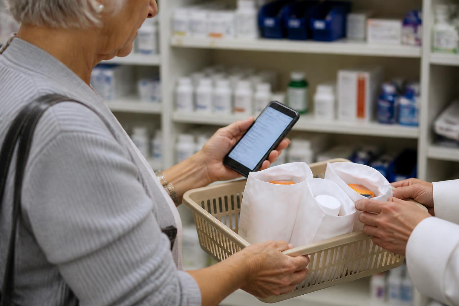 An older adult checking a medication list on a blurred phone while comparing generic prescription bags in a basket.