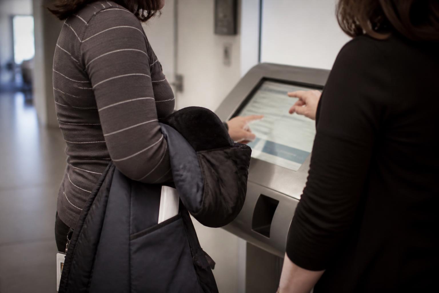 An employee adjusting a paycheck setting at the kiosk while a payroll staffer points from the side.
