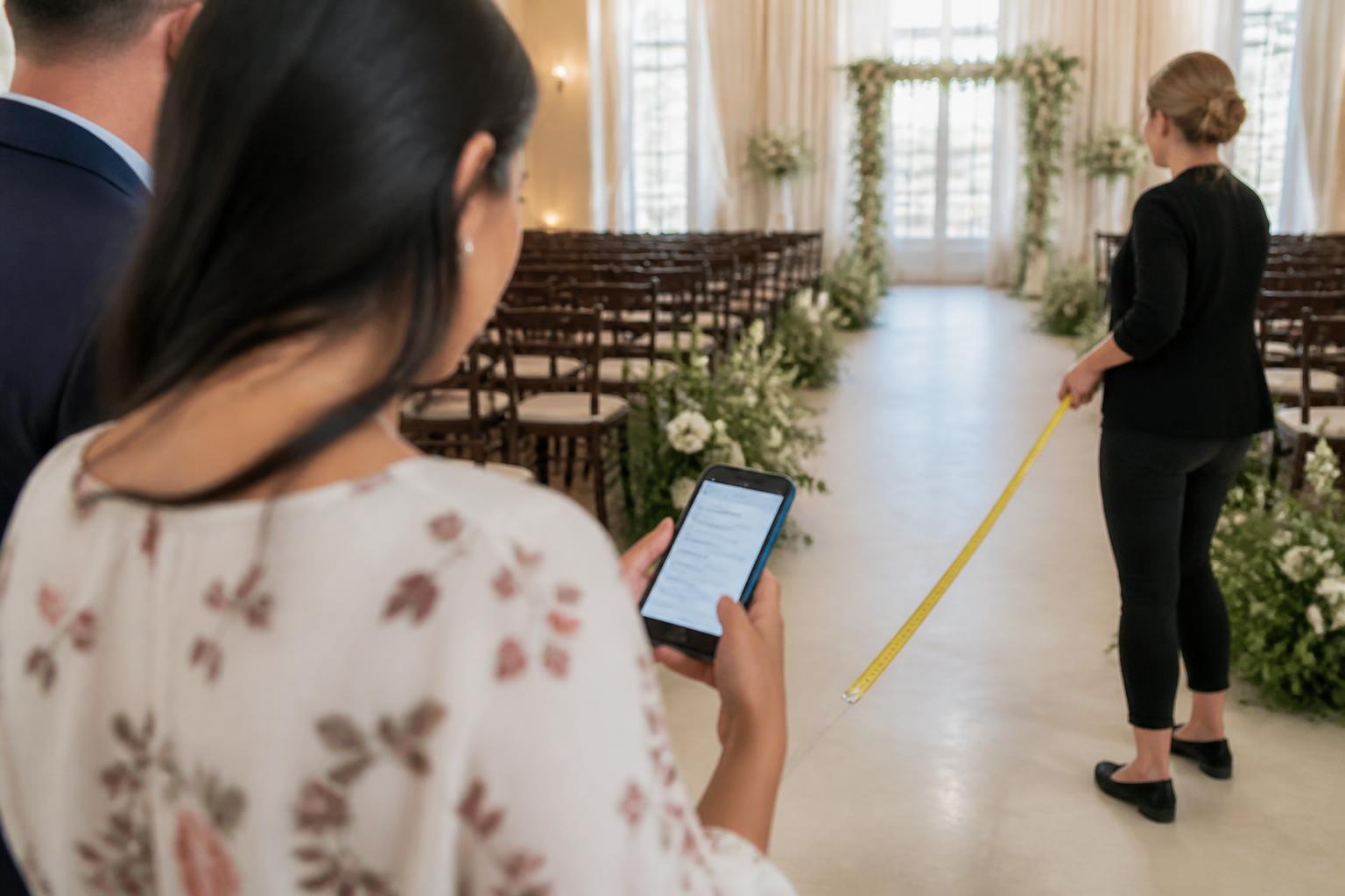 A couple measuring the space with a venue coordinator while one person checks a blurred budget note on a phone.